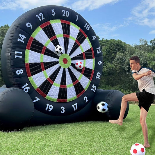 Person kicking a soccer ball towards a large inflatable dartboard on grass with trees in the background.